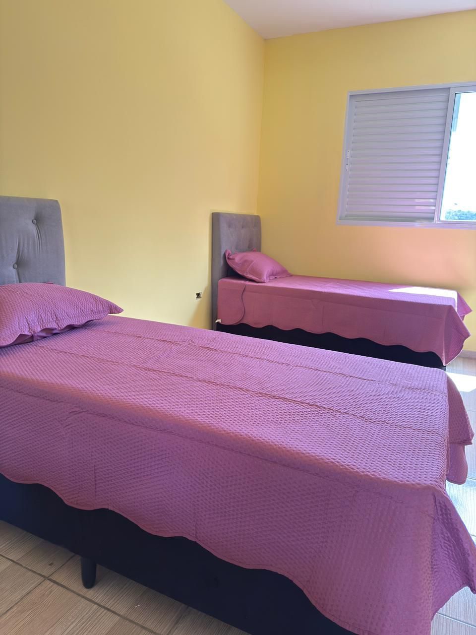Quarto feminino com cama arrumada em tons de rosa, poltrona e janela iluminada